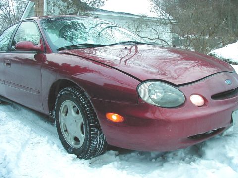 1997 Ford Taurus - A red 1997 Ford Taurus sits in the snow, its headlights on.