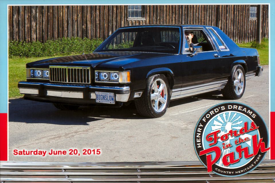 Black two-door Ford Panther sedan with custom wheels parked by a barn at a car show