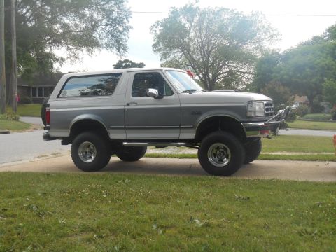 1994 Ford Bronco - A lifted, two-tone gray and white 1994 Ford Bronco is parked on a paved driveway with a grassy lawn in the foreground. The SUV has large of…