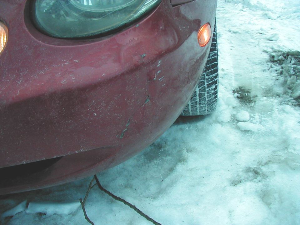 Close-up of a red car's front bumper and headlight in the snow. Tire visible