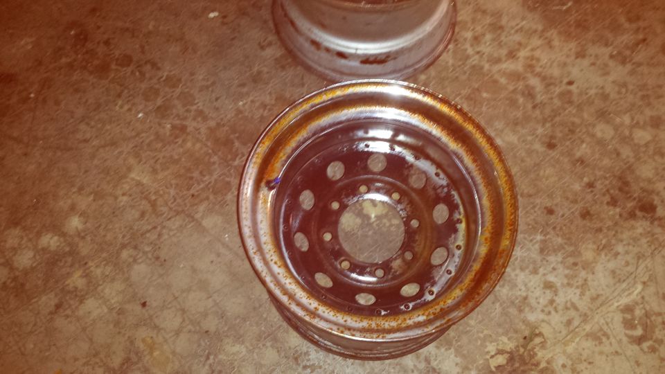 Two rusty steel wheels on a floor. One wheel is closer and shows bolt holes and a rusted rim.