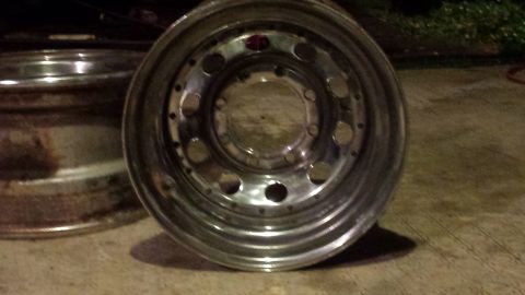 Polished metal wheel with circular cutouts and bolt holes, with a rusty wheel in the background.