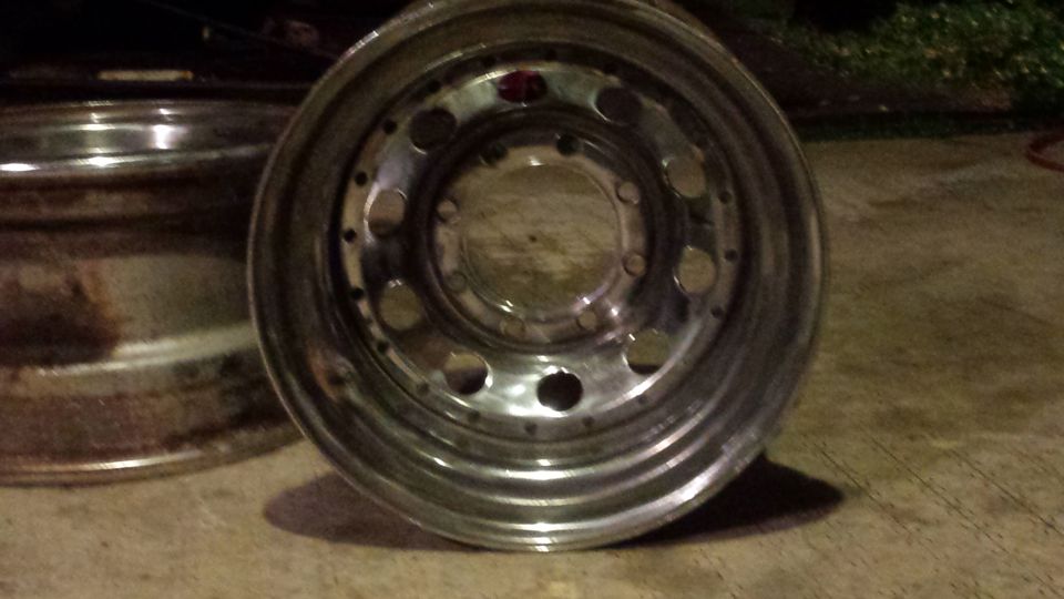 Polished metal wheel with circular cutouts and bolt holes, with a rusty wheel in the background.