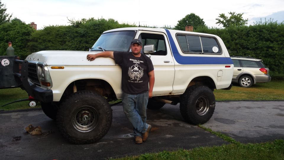 Man leaning on a lifted white 1979 Ford Bronco with a blue stripe, silver Subaru Outback in background.