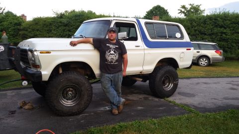 Lifted white 1970s Ford Bronco with blue stripe, custom bumper, and large tires, man leaning on it.