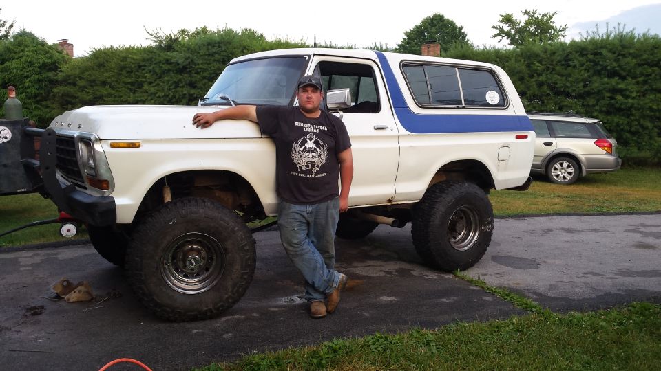 Lifted white 1970s Ford Bronco with blue stripe, custom bumper, and large tires, man leaning on it.
