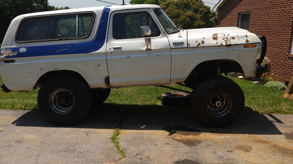 Lifted white 1979 Ford Bronco with blue stripe, showing rust and modifications.