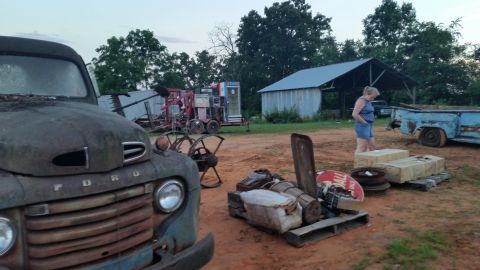 2015 Chevrolet new - A rusty, vintage Ford pickup truck is parked in a yard with other old vehicles and equipment. A woman is standing near some boxes and tires…