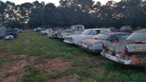 2015 Chevrolet new - A line of several old, rusty vehicles, mostly cars and trucks, are parked in a grassy field with trees in the background during dusk.