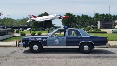 1991 Ford LTD Crown Victoria - Photograph of a Massachusetts State Police cruiser shown in profile with a military jet on display in the background.