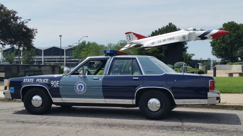 1991 Ford LTD Crown Victoria - Photograph of a Massachusetts State Police cruiser with a rooftop light bar in front of a fighter jet monument.