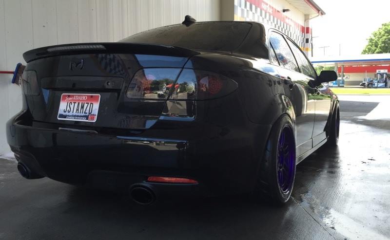 Black Mazda 6 sedan with tinted taillights, spoiler, and purple wheels, parked at a car wash.