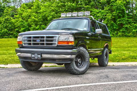 1995 Ford Bronco - A black Ford Bronco is parked on the side of a road with a grassy area and trees in the background. The vehicle has five auxiliary lights m…