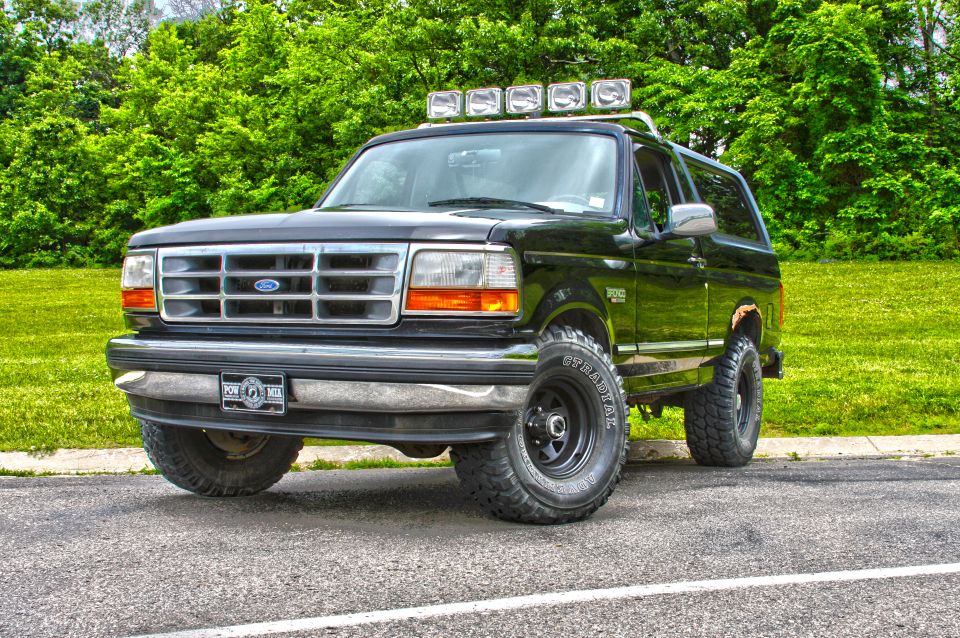 Black Ford Bronco with roof lights parked on asphalt with grass and trees behind.