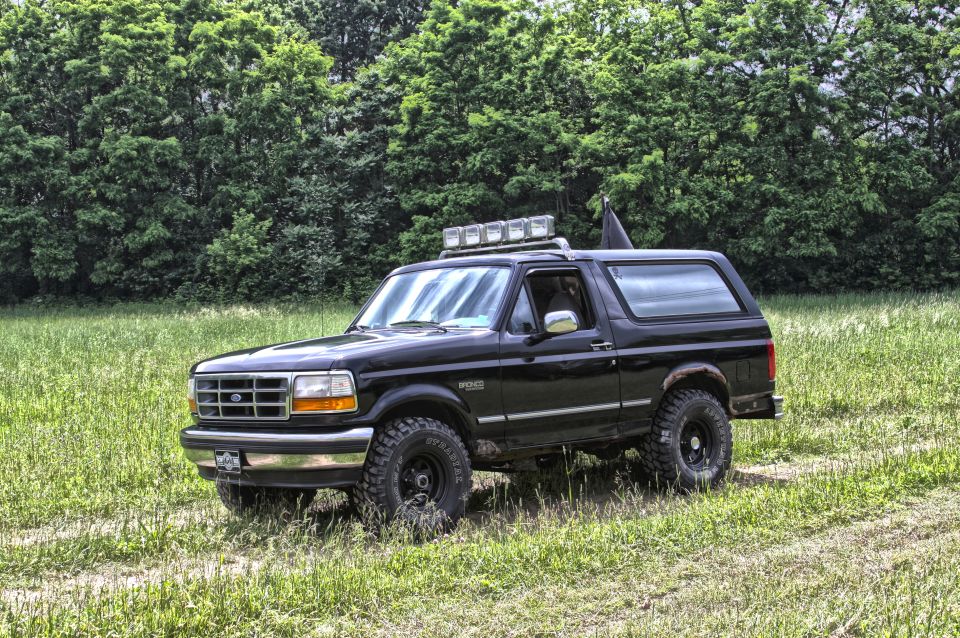Black 1995 Ford Bronco with roof rack lights and flag in a grassy field.