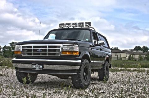 1995 Ford Bronco - A black Ford Bronco is parked on a gravel lot with a field and buildings in the background. The SUV has a light bar mounted on the roof.