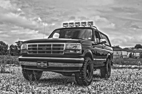 1995 Ford Bronco - A black Ford Bronco is parked on a rocky, grassy terrain under a cloudy sky. The SUV features a roof-mounted light bar and large off-road t…