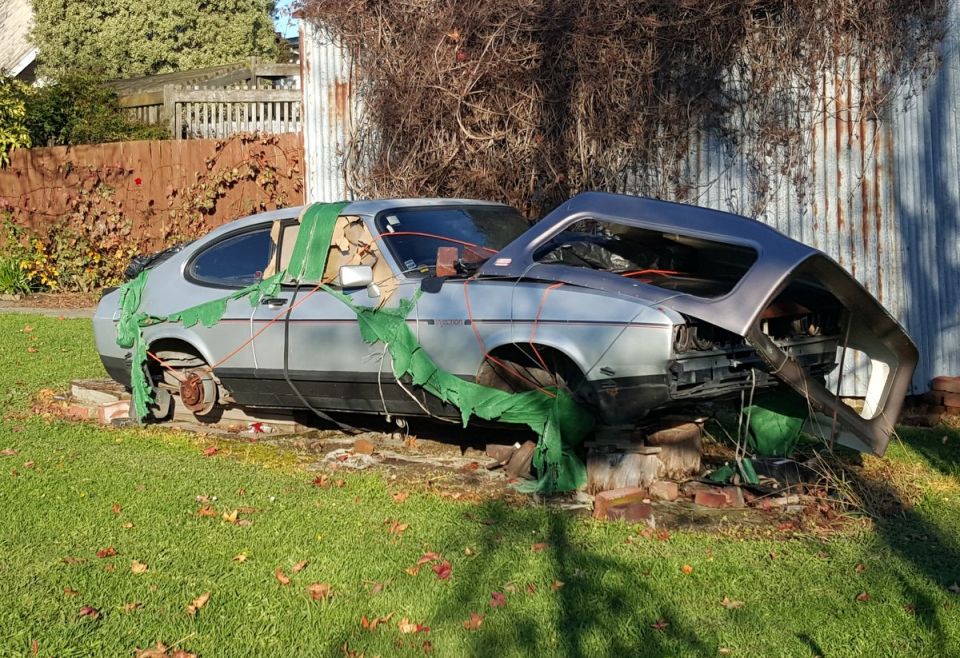 Damaged silver coupe with hood and roof removed, draped in green tarps.