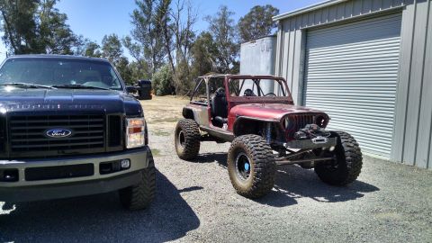 1985 Jeep CJ7 - A custom red Jeep CJ7 with large off-road tires and a roll cage sits parked next to a black Ford Super Duty pickup truck in front of a meta…