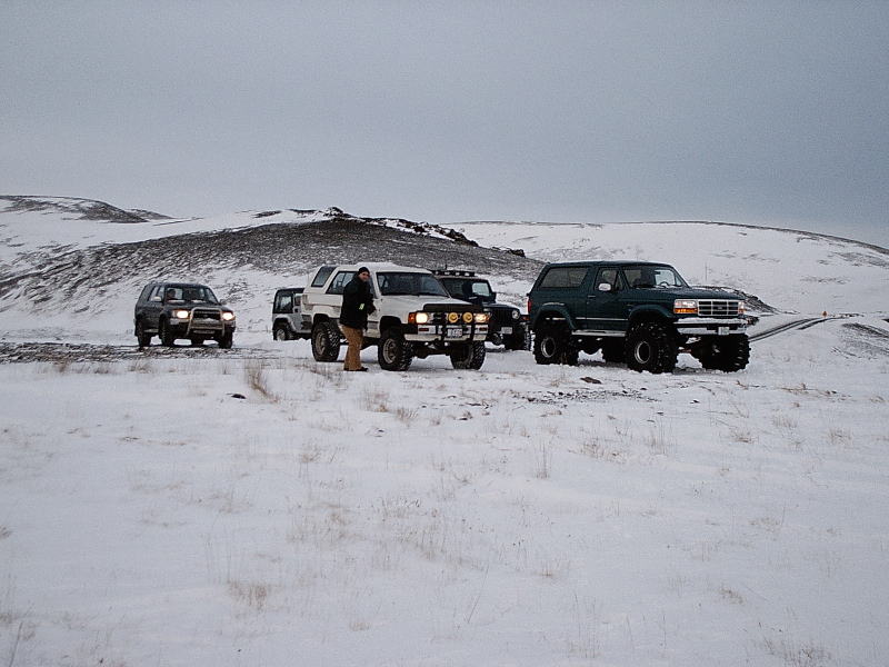 Green Ford Bronco and other lifted 4x4s parked on snowy hill under gray sky.