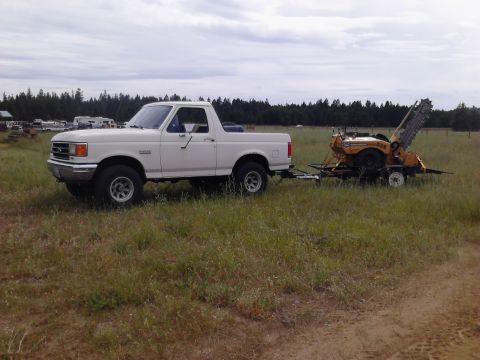 1990 Ford Bronco - A white 1990 Ford Bronco is parked in a grassy field, towing a yellow trencher on a trailer. The vehicle is viewed from the side, with a li…