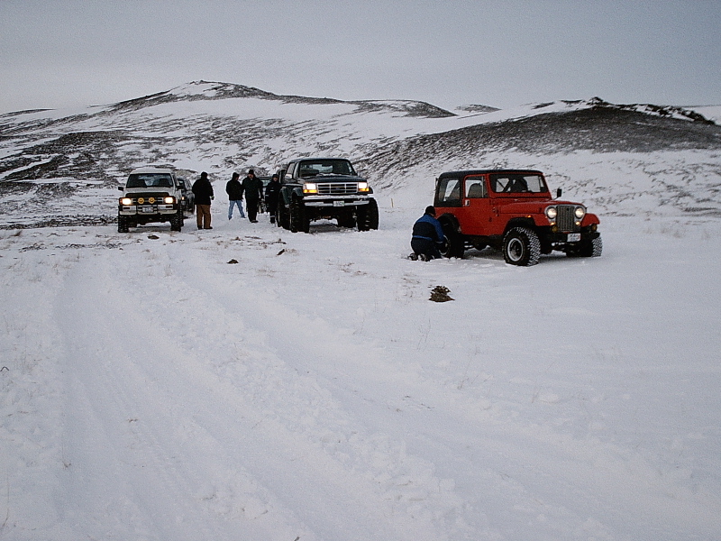 Group of 4x4 trucks and a red Jeep parked on snowy hills with people