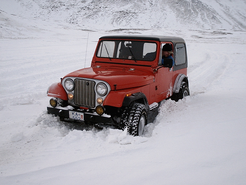 Red Jeep with black hardtop driving through deep snow, front three-quarter view