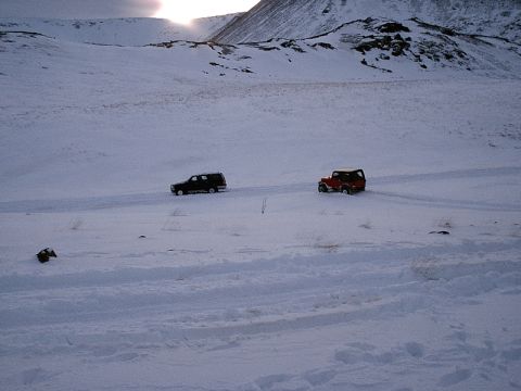 1996 Ford Bronco - Two SUVs traverse a snow-covered terrain under a cloudy sky with mountains in the distance.