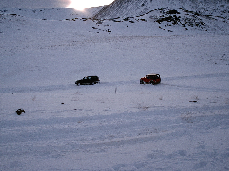 Dark SUV and red open-top 4x4 parked in a snowy mountain valley at dusk