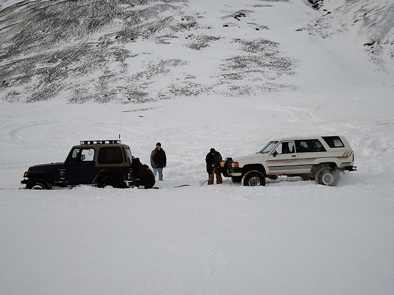 Dark Jeep and white SUV stuck in snow with people rigging a recovery line