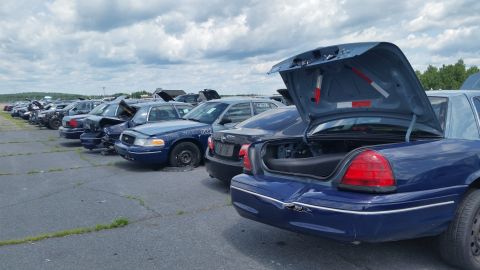 2001 Ford Crown Victoria - Junkyard scene with multiple Ford Crown Victoria police cars lined up under a cloudy sky.