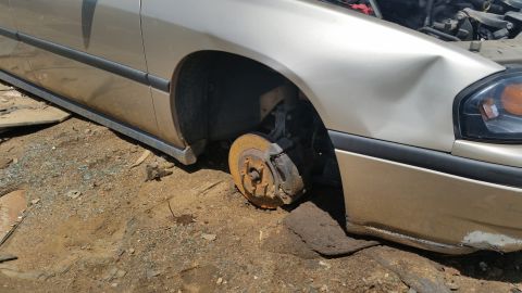 2001 Ford Crown Victoria - A gold sedan sits in a junkyard with its front passenger wheel removed and the fender damaged.