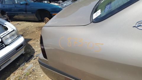 2001 Ford Crown Victoria - A tan sedan's rear quarter panel is marked with orange numbers in a junkyard setting with other vehicles visible.
