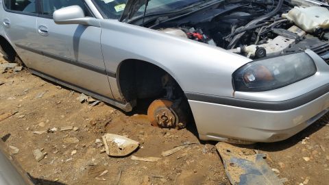 2001 Ford Crown Victoria - A damaged silver sedan sits in a junkyard with its hood open and front passenger wheel removed.