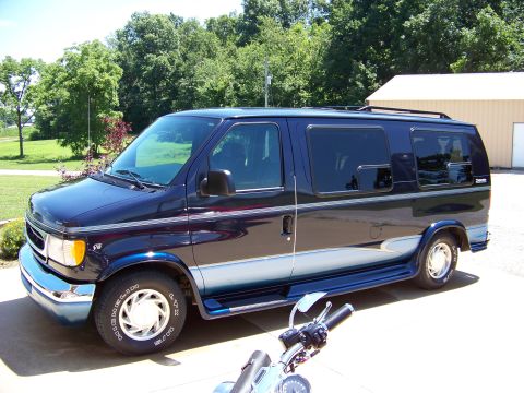 2000 Ford E150 - A dark blue Ford E-Series van is parked in front of a building with trees in the background. A motorcycle is partially visible.