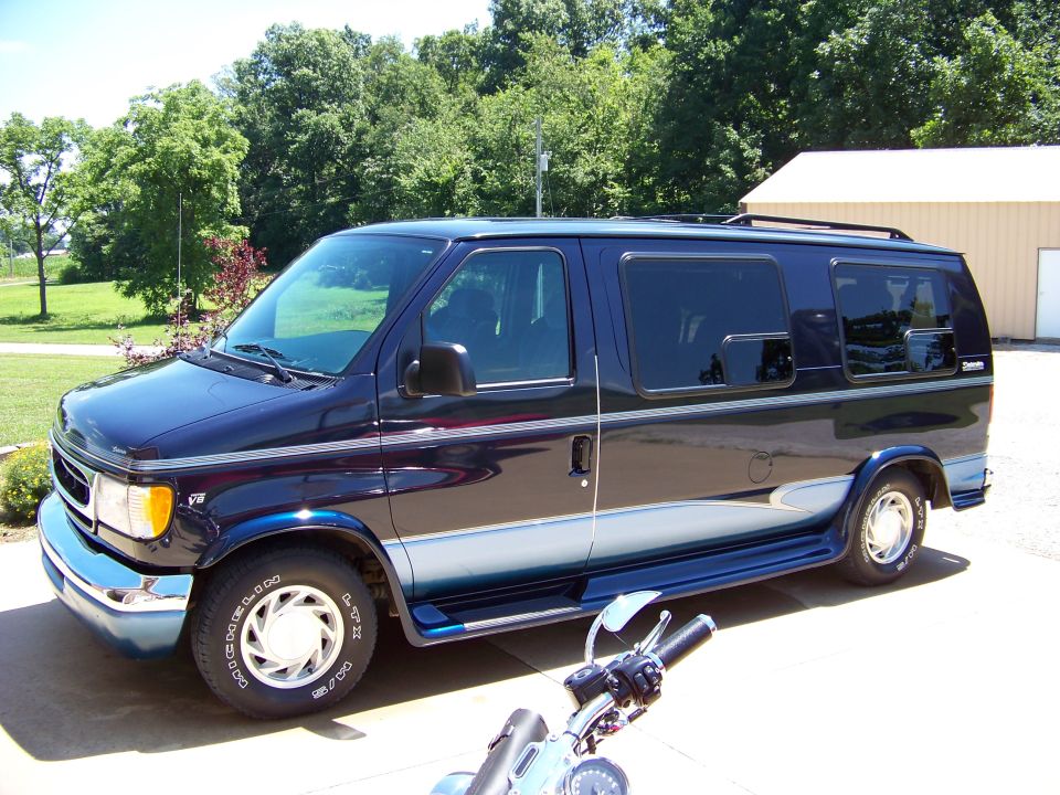 Dark blue Ford E-Series van parked outside a building. Motorcycle handlebars are in the foreground