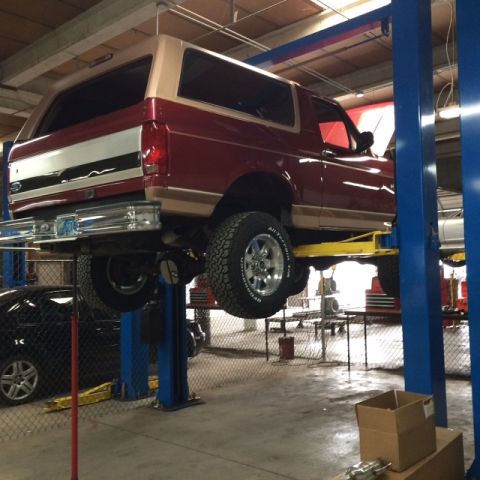 1994 Ford Bronco - A red and tan Ford Bronco is lifted on a two-post car lift in a garage. The rear of the SUV is visible, showing its tires and suspension co…