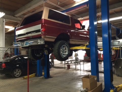 1994 Ford Bronco - A red and tan Ford Bronco is raised on a hydraulic lift in an auto repair shop. The rear of the SUV is visible, showing its tailgate and ch…