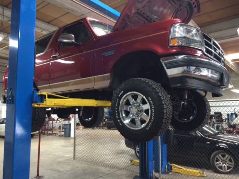 1994 Ford Bronco - A red Ford Bronco is raised on a hydraulic lift in a repair shop, with its hood open. The vehicle has off-road tires and chrome wheels.