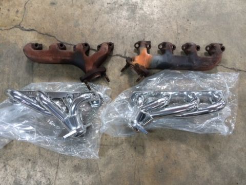 1994 Ford Bronco - Two sets of exhaust manifolds are laid out on a concrete floor. The set on the left is new and chrome-plated, still in plastic packaging. T…