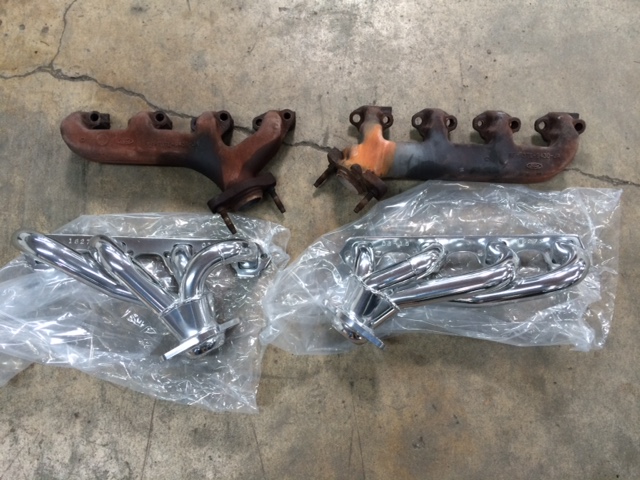 Two sets of exhaust manifolds, one new and chrome, one used and rusted, on a concrete floor.