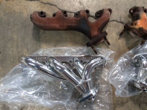 1994 Ford Bronco - Two sets of exhaust manifolds are laid out on a concrete surface. One set is new and chrome-plated, wrapped in plastic. The other set is ol…