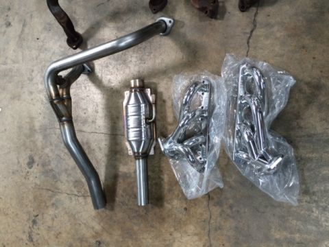 1994 Ford Bronco - Two chrome exhaust headers are packaged in plastic wrap, alongside a catalytic converter and exhaust pipes on a concrete floor.