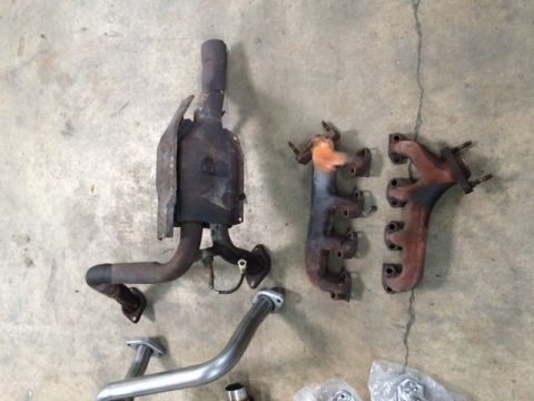 1994 Ford Bronco - Three exhaust manifolds and a catalytic converter are laid out on a concrete floor. The manifolds vary in size and color, with one showing…