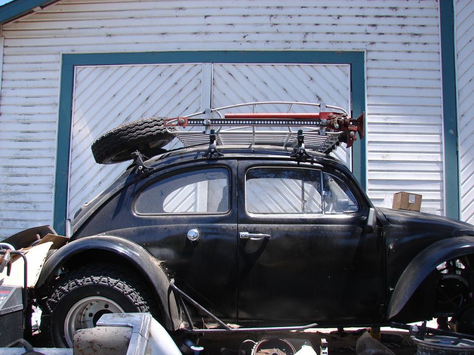 Black Volkswagen Beetle with roof rack, spare tire, and Hi-Lift jack in front of a white garage.