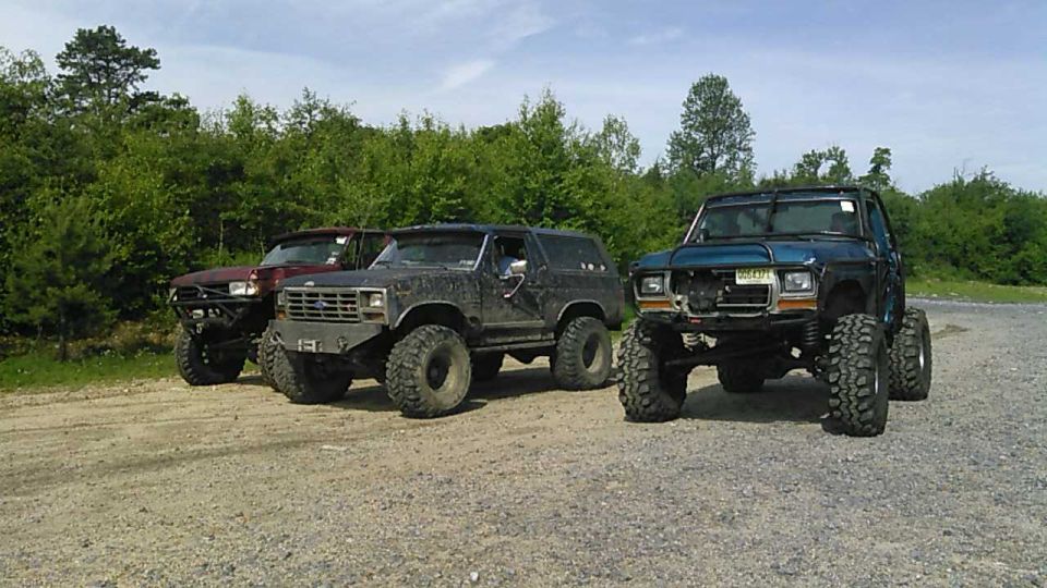 Three lifted SUVs, red, dark gray, and teal, parked on a gravel lot with trees in the background.