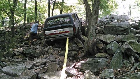 1988 Ford Bronco - A man in a blue shirt and shorts is assisting a black and silver SUV that is navigating a rocky, wooded trail. A yellow tow strap is attach…