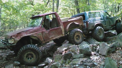 1988 Ford Bronco - Two off-road vehicles, a red pickup truck and a dark SUV, navigate a rocky, wooded trail. The red truck is tilted, with its driver's side d…