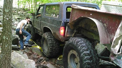 1988 Ford Bronco - A man in a white t-shirt and blue jeans is assisting a modified dark-colored Ford Bronco as it navigates a rocky, wooded terrain. The Bronc…