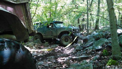 1988 Ford Bronco - A dark green, two-door SUV navigates a rocky, wooded trail. A yellow tow strap is attached to the front bumper.